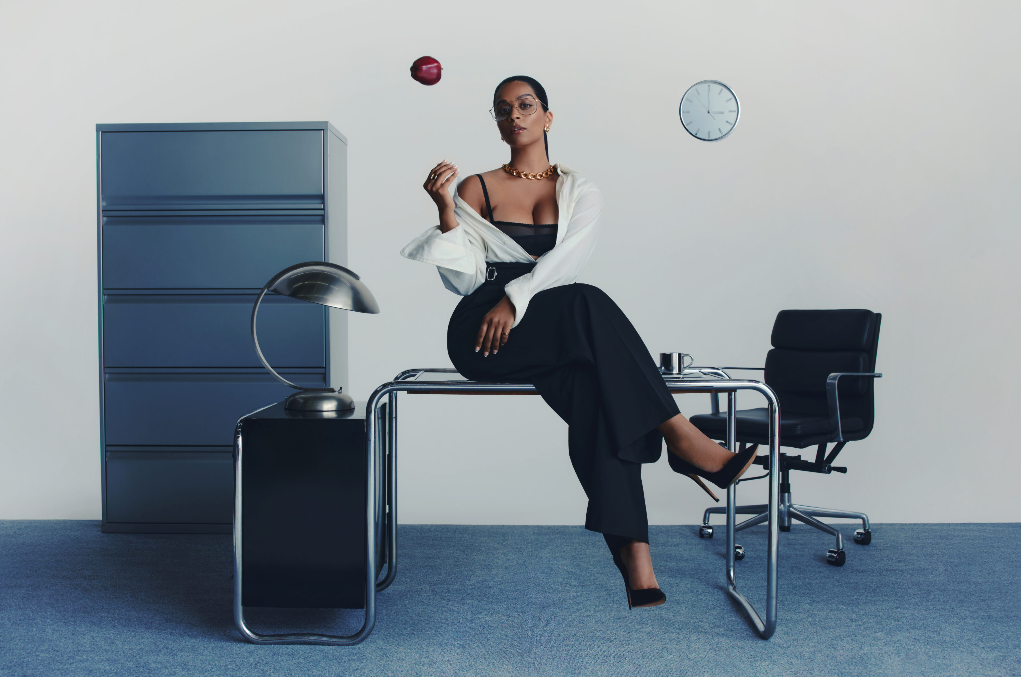 Writer-actress-producer Lilly Singh sits on a desk and tosses an apple in the air, eyes locked on the camera
