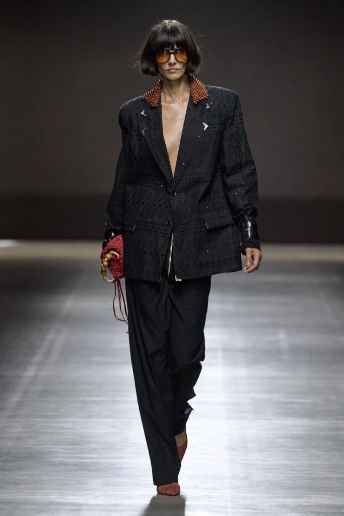 Model walking down the runway wearing a textured black blazer and black pants.