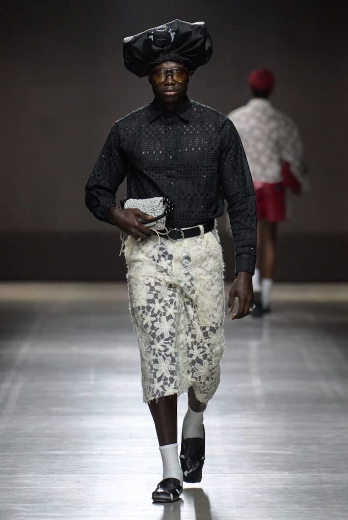 Male model walking down the runway wearing white lace pants and a textured black shirt.
