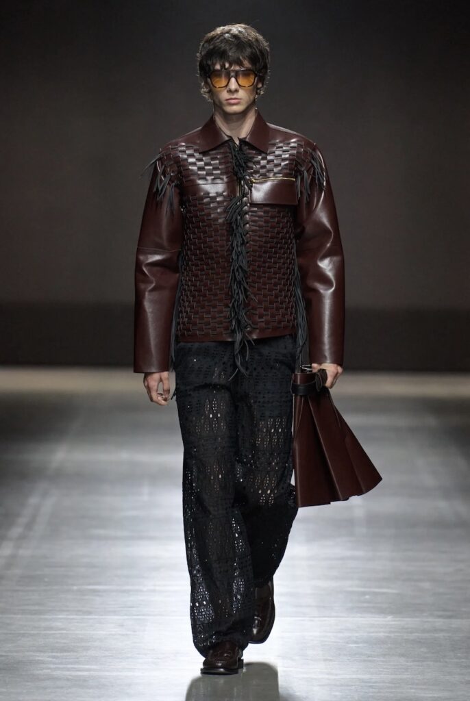 Male model walking down the runway in a brown leather jacket and textured black pants.