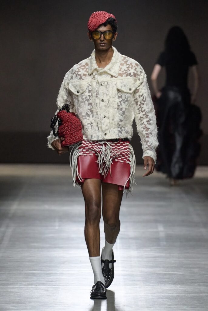 Male model walking down the runway with red shorts and a flower lace jacket.