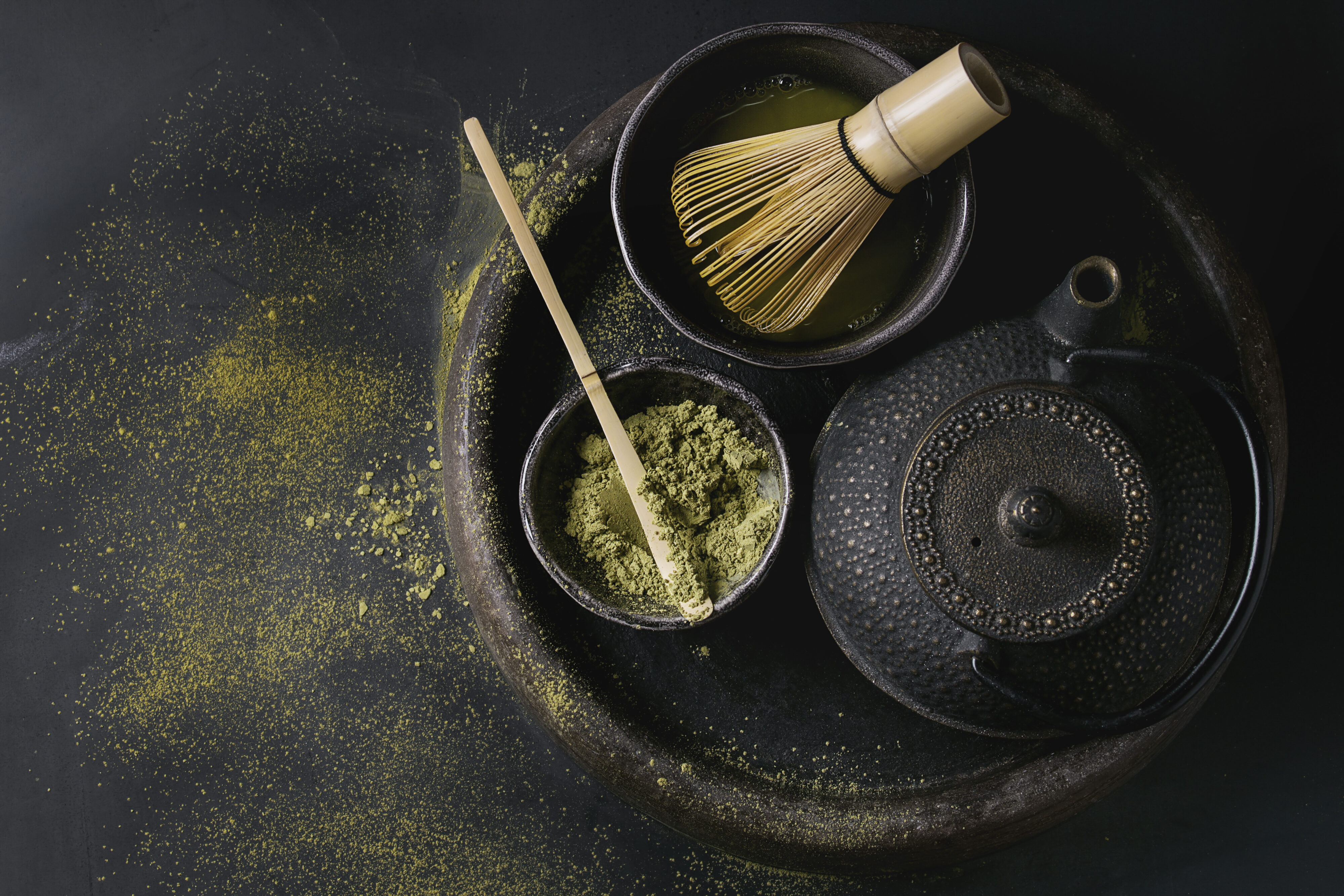 Green tea matcha powder and hot drink in black bowls standing with iron teapot and bamboo traditional whisk in terracotta tray over dark metal background.