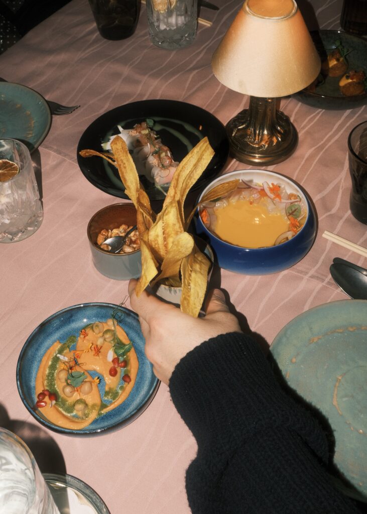 A person holds a cup of plantain chips near a cozy dining table with small plates of colourful dishes.