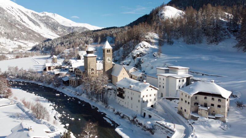 An alpine village covered in snow with a flowing river in the middle and mountains in the background.