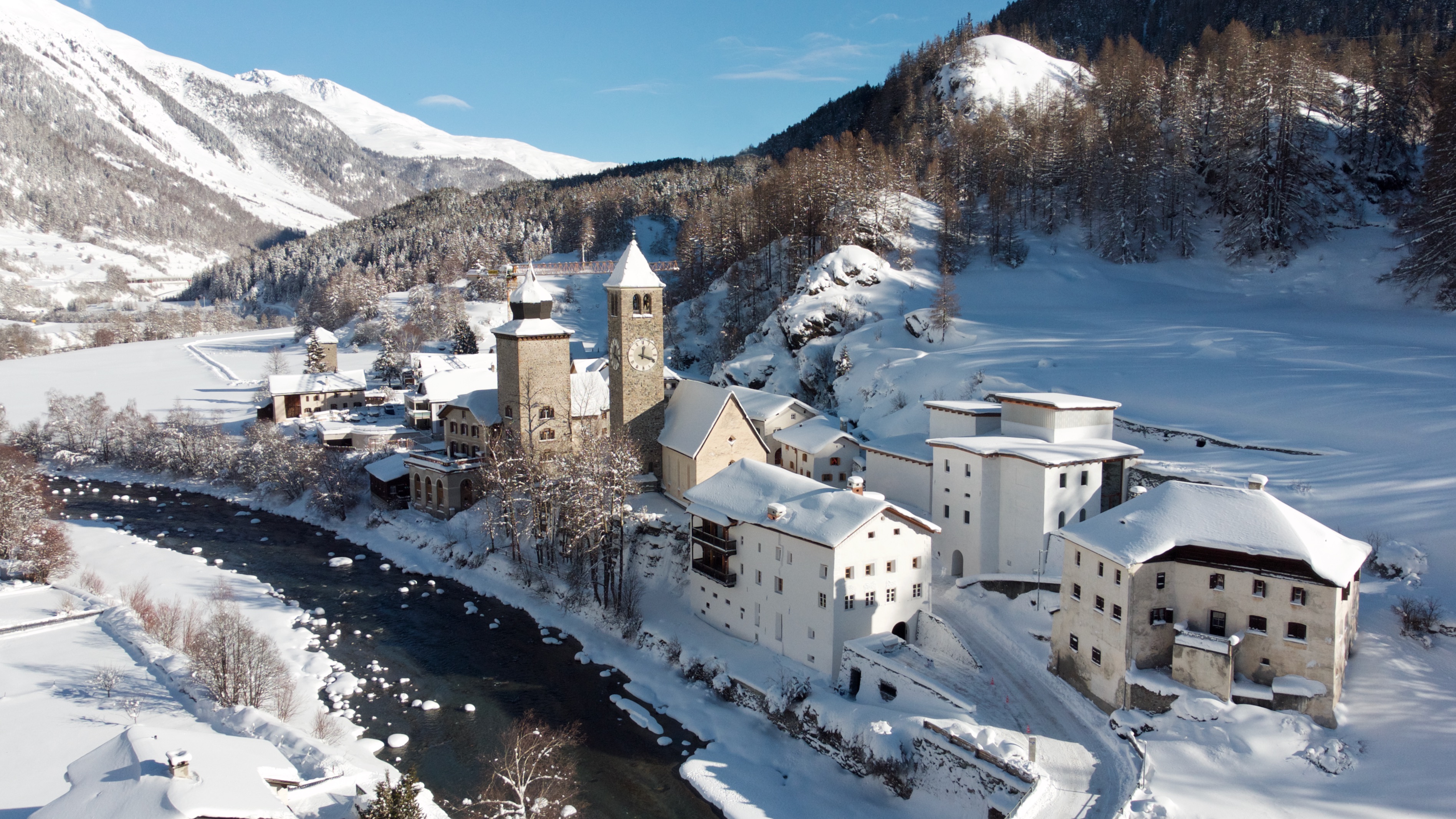 An alpine village covered in snow with a flowing river in the middle and mountains in the background.