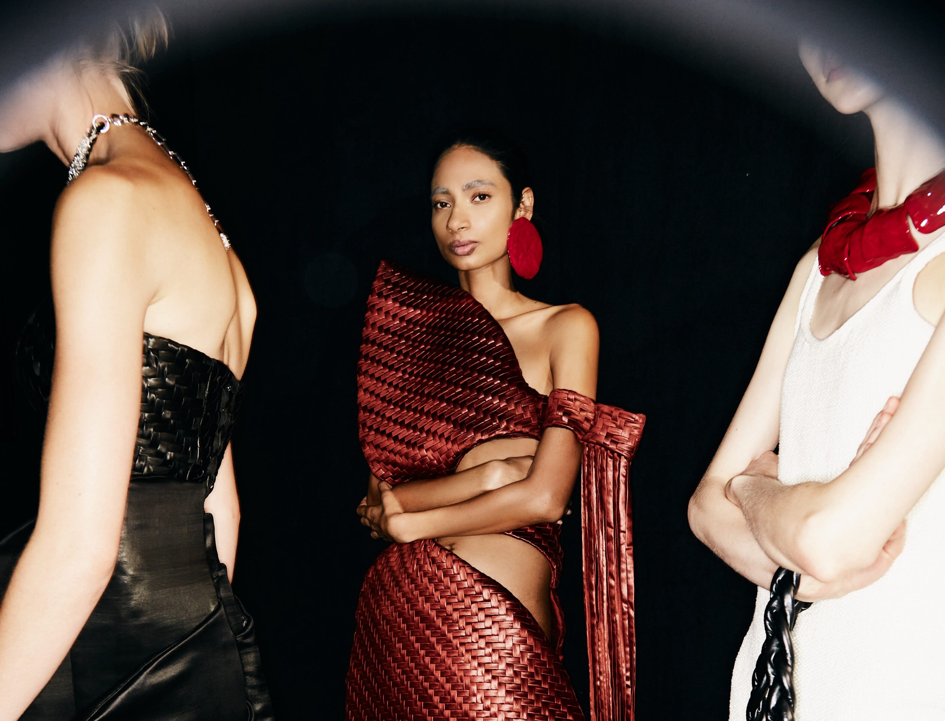 Three fashion models stand against a dark background. The model in the middle is facing forward and wearing a woven red dress.