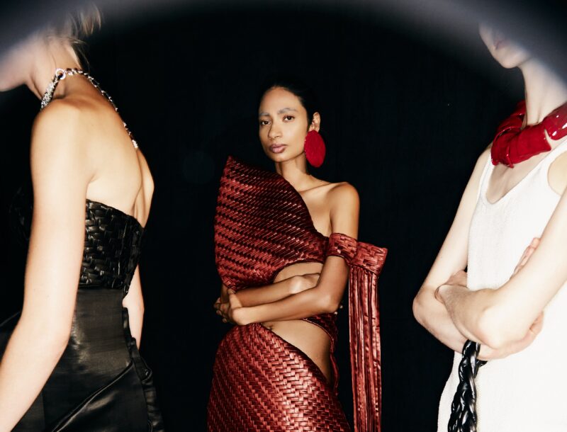 Three fashion models stand against a dark background. The model in the middle is facing forward and wearing a woven red dress.