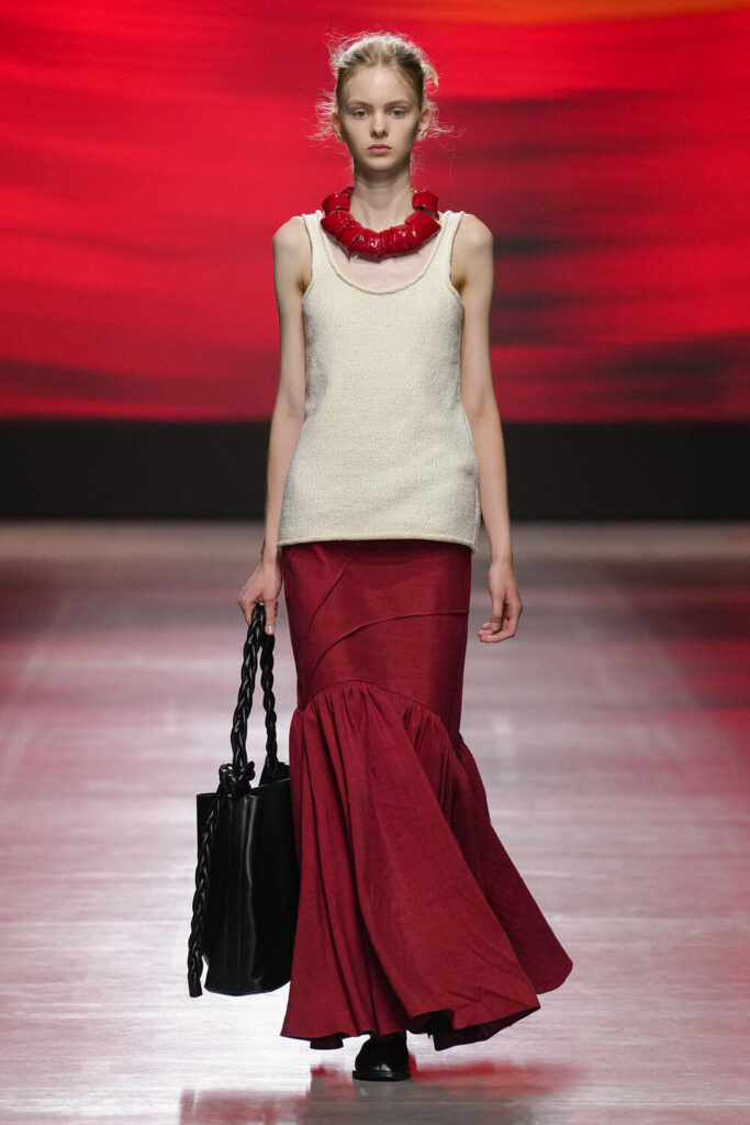 Female model walking down the runway wearing a long cream tank top, a chunky red necklace and a vibrant red skirt.