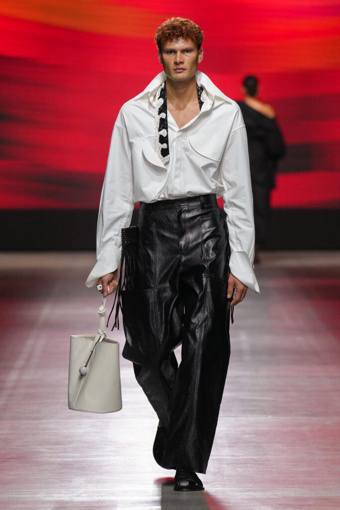 Male model walking down the runway in a white long sleeve dress shirt and black leather pants.