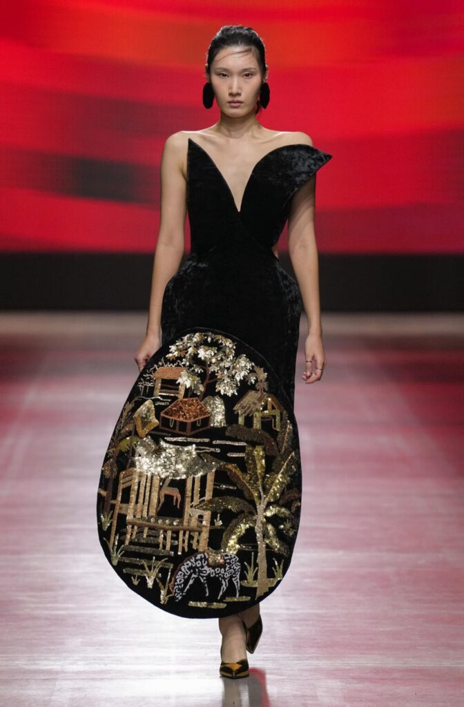 Female model walking down the runway in a black velvet dress with a sequinned gold embellishment that has village and nature imagery.