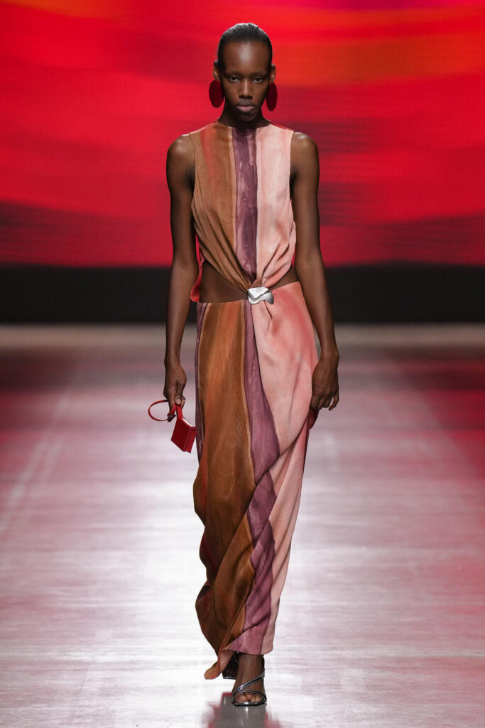 Female model walking down the runway wearing a long dress with burnt orange, dark purple and light pink colour tones.