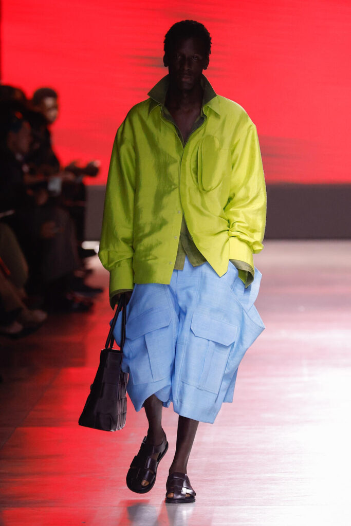 Male model walking down the runway wearing a neon yellow jacket and oversized sky blue shorts.