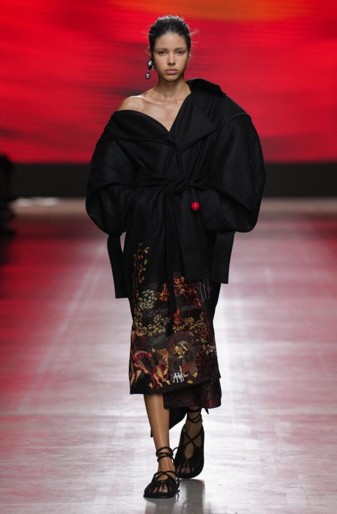 Female model walking down the runway wearing an oversized black jacket with a printed skirt.